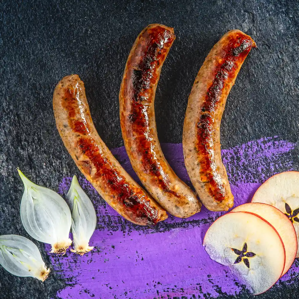 gebratene Bratwurst auf lila Hintergrund umgeben von Apfel und Zwiebeln