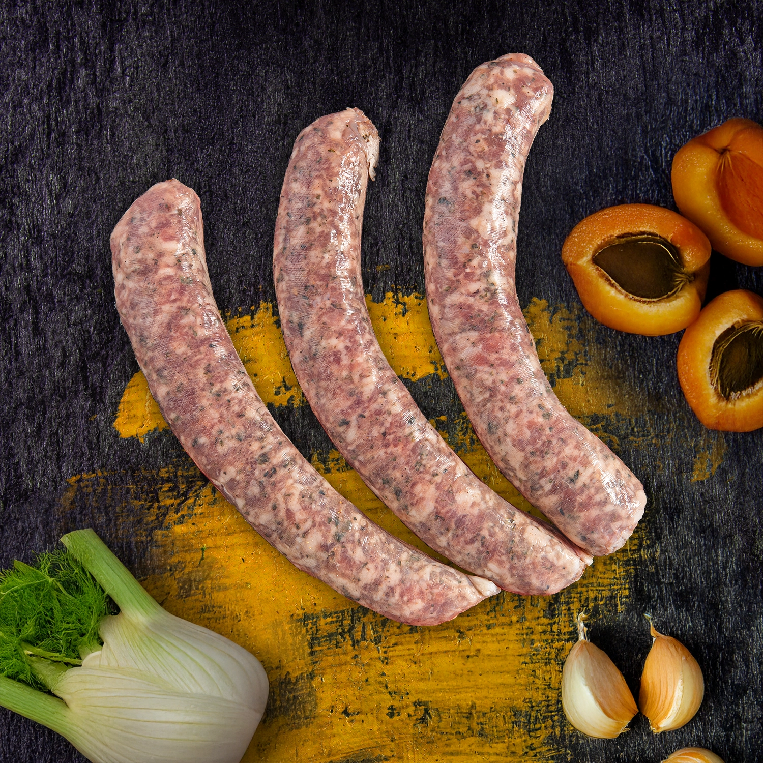 Drei rohe Bratwürste neben Fenchelknolle, Knoblauchzehen und Aprikosenhälften.