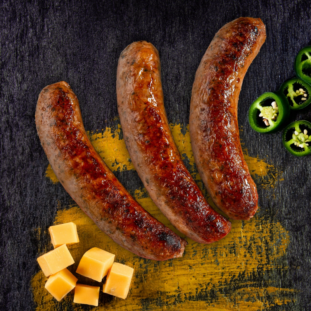 Drei gebratene Bratwürste neben Jalapeno Scheiben und Cheddar Würfeln.