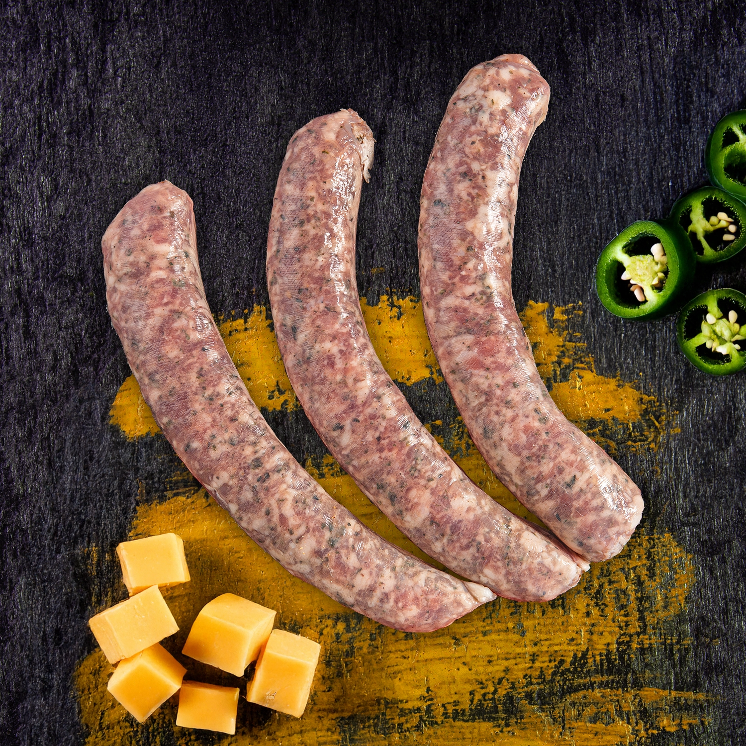 Drei rohe Bratwürste neben Jalapeno Scheiben und Cheddar Würfeln.